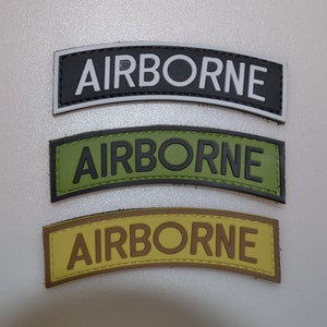May include: Three curved patches with the word "AIRBORNE" in bold letters. The top patch is black with white text, the middle is olive green with black text, and the bottom is tan with brown text. The patches are likely military or tactical.