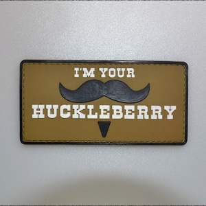 Puede incluir: Un parche rectangular con el texto "I'M YOUR HUCKLEBERRY" en letras mayúsculas blancas. Un bigote negro está encima de la palabra "HUCKLEBERRY". El parche tiene un fondo marrón y un borde negro.