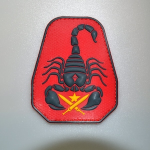 May include: A red patch with a black scorpion design. The patch has a textured surface and a black border. Below the scorpion are two crossed yellow swords and a yellow star.