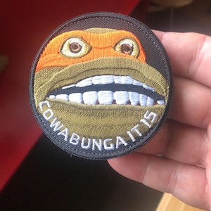 Puede incluir: Parche redondo bordado con la cara de una Tortuga Ninja, con máscara naranja y una amplia sonrisa. El texto "COWABUNGA IT IS" está bordado alrededor del borde del parche.
