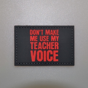 Può includere: Una toppa rettangolare nera con la scritta "DON'T MAKE ME USE MY TEACHER VOICE" in rosso. La toppa ha un bordo cucito ed è probabilmente progettata per essere attaccata a vestiti o borse. Lo sfondo è grigio chiaro.
