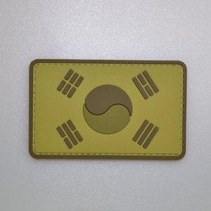May include: A rectangular patch featuring the South Korean flag design. The patch has a light yellow background with a brown border. The flag's symbols are in brown and the center emblem is a lighter brown.
