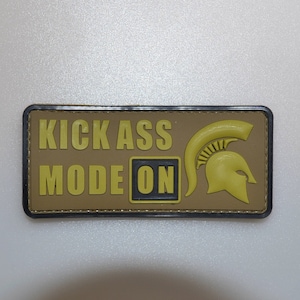 Puede incluir: Un parche rectangular con el texto "KICK ASS MODE ON" en letras amarillas. Un casco espartano amarillo está en el lado derecho. El parche tiene un fondo marrón y un borde negro.