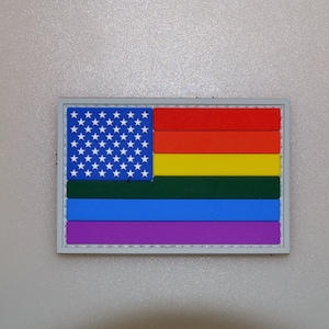 May include: A rectangular patch combining the American flag and the rainbow pride flag. The upper left corner shows the blue field with white stars, and the rest of the flag has horizontal stripes in red, orange, yellow, green, blue, and purple.