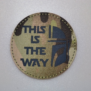 Könnte beinhalten: Runder Aufnäher mit Tarnmuster und dem Text "THIS IS THE WAY" in Dunkelgrau. Eine dunkelgraue Helm-Grafik befindet sich auf der rechten Seite. Der Aufnäher hat einen genähten Rand.