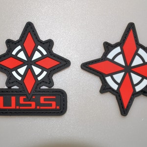 Könnte beinhalten: Zwei schwarz, rot und weiß gestickte Aufnäher. Ein Aufnäher zeigt ein Kompassrosen-Design mit dem Text "U.S.S." in Rot. Der andere Aufnäher ist ein Kompassrosen-Design. Beide Aufnäher haben einen schwarzen Rand.