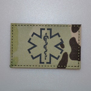 May include: A rectangular patch with a camouflage pattern and a black medical symbol. The symbol features a staff with a snake entwined around it, set within a six-pointed star. The patch is bordered by black stitching.