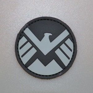 May include: A round patch featuring the S.H.I.E.L.D. logo. The patch has a black border and a gray and black emblem of a stylized eagle. The patch is likely made of a durable material.