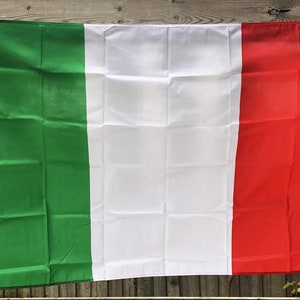 Puede incluir: Bandera italiana con franjas verticales verdes, blancas y rojas. La bandera está hecha de una tela ligera y está colgada al aire libre. Es rectangular y tiene ojales a lo largo del borde izquierdo para colgarla.