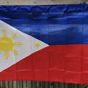 Puede incluir: Bandera rectangular de Filipinas, con una sección superior azul y una inferior roja. Un triángulo blanco en el lado del mástil contiene un sol amarillo con ocho rayos y tres estrellas amarillas. La bandera es de tela brillante.