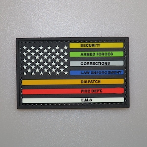 Puede incluir: Un parche rectangular con un diseño estilizado de la bandera estadounidense. Las estrellas son blancas sobre un fondo negro. Las franjas de colores representan diferentes servicios: seguridad, fuerzas armadas, correcciones, policía, despacho, bomberos y E.M.S.