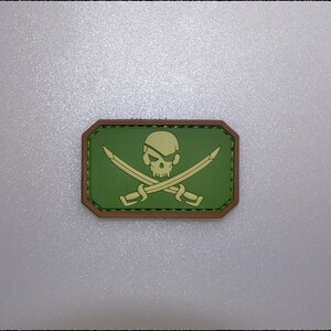 Könnte beinhalten: Ein rechteckiger Aufnäher mit Piratenmotiv. Er zeigt einen Totenkopf mit Augenklappe und gekreuzten Schwertern in Hellgrün und Beige auf einem dunkelgrünen Hintergrund mit braunem Rand. Für Kleidung geeignet.