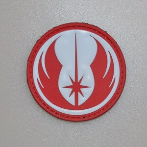 Puede incluir: Parche redondo rojo y blanco con un emblema estilizado. El diseño incluye una forma blanca similar a un pájaro y una estrella en el centro, sobre un fondo rojo. El parche tiene un borde cosido.