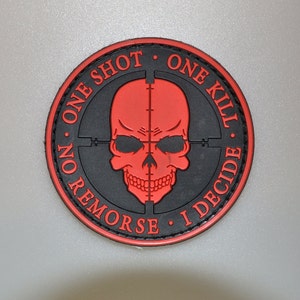 Pode incluir: Remendo redondo preto e vermelho com uma caveira vermelha e uma mira. O texto "ONE SHOT, ONE KILL, NO REMORSE, I DECIDE" está impresso ao redor da caveira. O remendo tem uma borda preta.