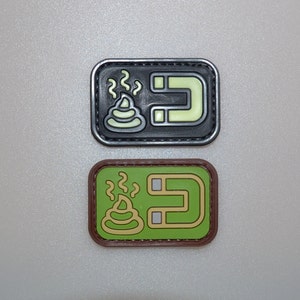 Könnte beinhalten: Zwei rechteckige Aufnäher mit einem humorvollen Design. Der obere Aufnäher ist schwarz mit einem leuchtend grünen Kackhaufen-Emoji und einem leuchtend grünen Magnet-Symbol. Der untere Aufnäher ist grün mit braunem Rand.