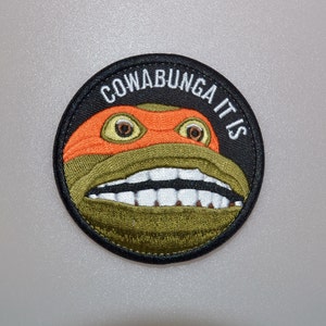 May include: Embroidered circular patch depicting a cartoon turtle's face with an orange mask and a wide grin. The patch features the words "COWABUNGA IT IS" in white on a black background, with a black border.