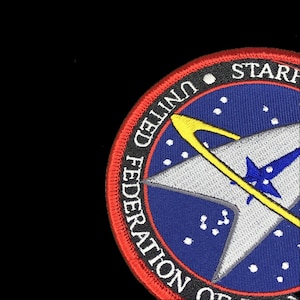 Peut inclure: Écusson brodé représentant l'emblème de Starfleet de Star Trek. L'écusson a un fond bleu avec une étoile blanche et des accents jaunes. Les mots "UNITED FEDERATION OF" et "STARF" sont brodés sur le bord en blanc.