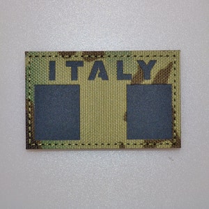 Pode incluir: Um patch retangular com a palavra "ITALY" em letras pretas. O patch tem um padrão de camuflagem em tons de verde, castanho e bege. Dois retângulos cinza escuro estão abaixo da palavra "ITALY". O patch é contornado com uma costura verde escura.