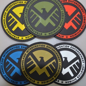 Op de afbeelding: Een verzameling ronde patches in verschillende kleuren, waaronder geel, groen, rood, blauw, goud en wit. Elke patch heeft een zwarte rand en een gestileerd adelaarslogo met de tekst "STRATEGIC HOMELAND INTERVENTION ENFORCEMENT LOGISTICS DIVISION".