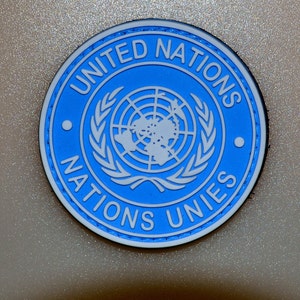 Pode incluir: Um patch redondo azul com as palavras "UNITED NATIONS" e "NATIONS UNIES" em branco. O centro apresenta um mapa-múndi branco rodeado por ramos de oliveira. Uma borda branca circunda o círculo azul.