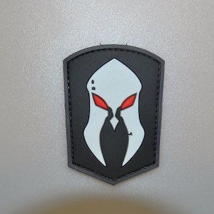 Może przedstawiać: Czarna i biała naszywka z wystylizowanym wzorem hełmu z czerwonymi oczami. Naszywka ma czarną obwódkę i kształt tarczy. Hełm jest biały z czerwonymi oczami.