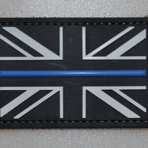 May include: A rectangular patch featuring the Union Jack flag design with a blue stripe across the center. The flag's white and gray elements are set against a black background. The patch has a black border with stitched detailing.