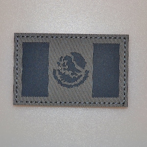 May include: A rectangular patch with the Mexican coat of arms. The patch has a textured, grey background with a dark grey emblem in the centre and two dark grey vertical rectangles. The edges are outlined with a blue stitch.