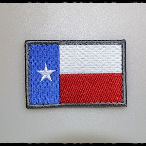 May include: Embroidered patch featuring the Texas state flag. The patch has a blue vertical stripe with a white star, a white horizontal stripe, and a red horizontal stripe. The patch is rectangular with a black border.