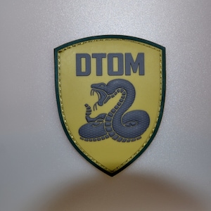 Puede incluir: Un parche en forma de escudo con fondo amarillo y borde verde oscuro. El parche presenta el texto "DTOM" en letras grises sobre una serpiente gris con la boca abierta.