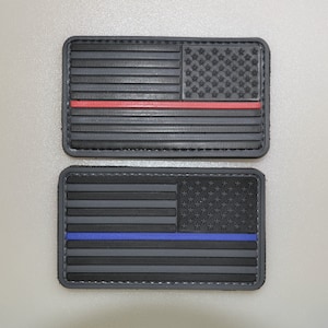 Könnte beinhalten: Zwei rechteckige Aufnäher mit dem Design der amerikanischen Flagge. Einer hat einen roten Streifen, der andere einen blauen Streifen. Beide sind hauptsächlich schwarz mit strukturierter Oberfläche und Rand.