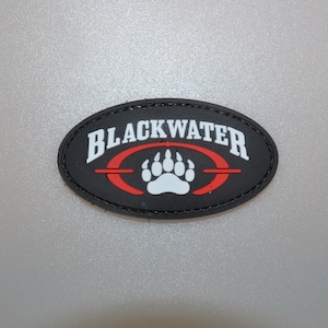 Op de afbeelding: Zwart ovaalvormig embleem met het woord "BLACKWATER" in witte letters. Een rood ovaal met een witte pootafdruk is eronder gecentreerd. De patch heeft een zwarte rand op een grijze achtergrond.