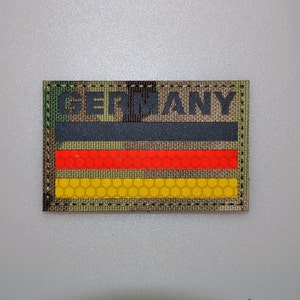 Könnte beinhalten: Ein rechteckiger Aufnäher mit dem Wort "GERMANY" in schwarzen Blockbuchstaben über einem schwarzen Streifen, einem roten Streifen und einem gelben Streifen. Die Streifen haben ein Wabenmuster. Der Aufnäher hat einen Tarnhintergrund.