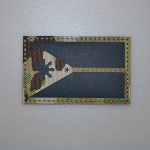 Pode incluir: Um patch retangular com um desenho de bandeira. O patch tem um fundo azul escuro com um triângulo bege e camuflado e uma faixa bege. O triângulo contém uma estrela e estrelas.