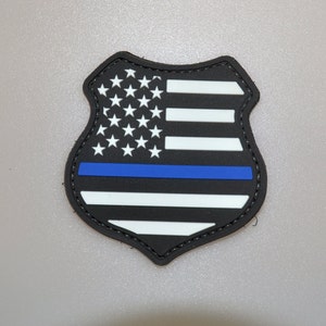 Puede incluir: Parche negro y blanco en forma de escudo con el diseño de la bandera estadounidense y una franja azul. El parche tiene un borde cosido. Las estrellas son blancas y las rayas se alternan entre blanco y negro. El fondo es gris.