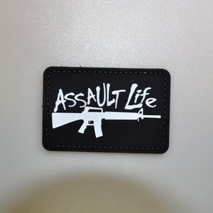 Könnte beinhalten: Schwarzer rechteckiger Aufnäher mit dem weißen Schriftzug "Assault Life" und einer weißen Gewehrgrafik. Der Aufnäher hat einen genähten Rand. Der Hintergrund ist hellgrau.