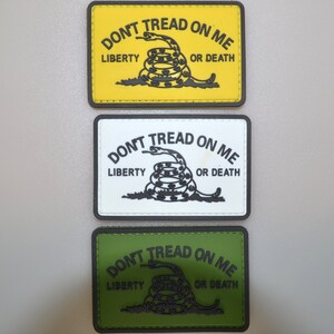 Può includere: Tre toppe rettangolari con la scritta "DON'T TREAD ON ME", "LIBERTY" e "OR DEATH" che circondano un serpente arrotolato. Le toppe sono gialle, bianche e verdi, ognuna con testo nero e un disegno di serpente nero.