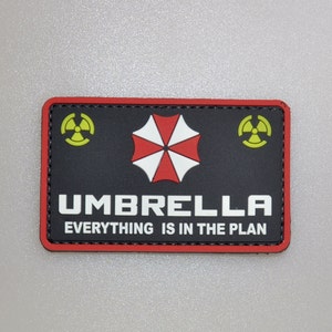 Puede incluir: Parche rectangular negro con borde rojo. El parche presenta la palabra "UMBRELLA" en letras mayúsculas blancas, un logotipo de paraguas rojo y blanco y el texto "EVERYTHING IS IN THE PLAN". Dos símbolos de peligro biológico amarillos están en el parche.