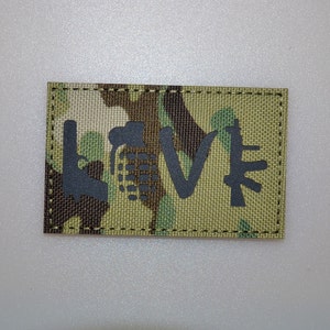 Può includere: Una toppa rettangolare con un motivo mimetico. La parola "LOVE" è scritta in nero, con ogni lettera stilizzata come un'arma: una pistola, una granata, un simbolo a forma di V e un fucile.