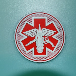 May include: A circular medical emblem in red and white, displaying the Star of Life symbol. It has a white border with a textured red outline. The central design features a staff with a snake, wings, and medical tools.
