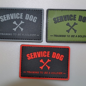 Pode incluir: Três patches retangulares com as palavras "SERVICE DOG" e "TRAINING TO BE A SOLDIER". Cada patch apresenta um gráfico de ossos cruzados. Os patches são cinza, verde-oliva e preto com letras vermelhas.