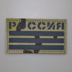 Pode incluir: Um patch retangular com a palavra cirílica "РОССИЯ" em letras de estêncil. O patch tem um fundo de camuflagem com três listras horizontais cinza. As bordas são costuradas com linha preta.