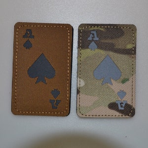 May include: Two rectangular patches with the Ace of Spades design. One is brown with a black spade symbol and the letter "A". The other has a camouflage pattern with a grey spade symbol and the letter "A".