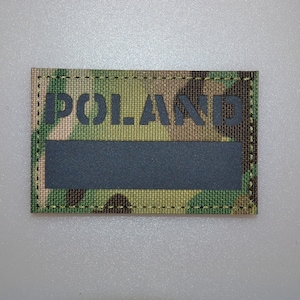 May include: A rectangular patch with the word "POLAND" in black block letters above a black rectangular area. The patch has a camouflage pattern in shades of green, brown, and tan. The edges are stitched with dark green thread.