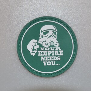 Puede incluir: Un parche redondo verde con borde blanco y el texto "YOUR EMPIRE NEEDS YOU...". Presenta una ilustración blanca de un casco de Stormtrooper y una mano señalando.