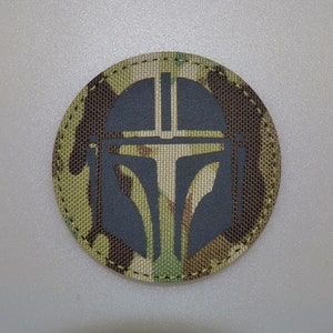 Könnte beinhalten: Ein runder Aufnäher mit einem Mandalorianer-Helm-Design. Der Hintergrund ist Tarnmuster in Grün-, Braun- und Beigetönen. Der Helm ist dunkelgrau. Der Aufnäher ist wahrscheinlich aus Stoff und hat einen genähten Rand. Der Durchmesser beträgt ca. 7,6 cm.