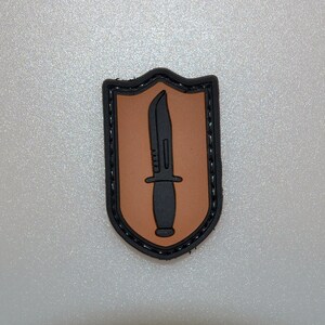 Könnte beinhalten: Ein schwarz-brauner Aufnäher mit einem schwarzen Messer auf braunem Hintergrund, umrahmt von einem schwarzen Rand. Der Aufnäher hat eine Schildform und ist wahrscheinlich ein Emblem oder eine Insignie.