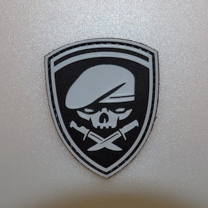 May include: A black and gray patch with a skull wearing a beret and crossed knives. The patch is shield-shaped with a gray skull and beret design on a black background. The patch has a gray border.