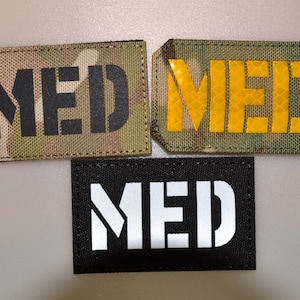 Könnte beinhalten: Drei rechteckige Aufnäher mit dem Wort "MED" in Großbuchstaben. Einer hat ein Tarnmuster, ein anderer ein reflektierendes gelbes Design, und der dritte ist schwarz mit weißen Buchstaben. Diese Aufnäher sind wahrscheinlich für medizinische oder taktische Zwecke.