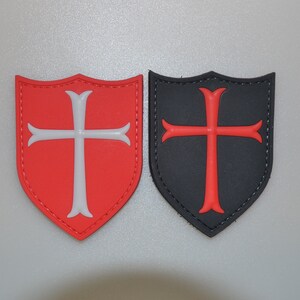 May include: Two shield-shaped patches. One is red with a white cross, the other is black with a red cross. Both have a stitched border. The patches are likely for clothing or accessories, featuring a heraldic design.