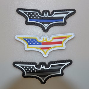 May include: Three patches in the shape of a bat symbol. The top patch has a blue line, the middle patch has the American flag design, and the bottom patch has a gray and black design. All patches have a black border.